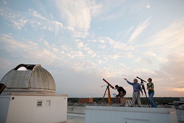 Three people on a roof top with telescopes observing the sky